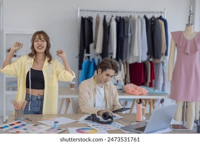 Two fashion designers enjoying creative workspace, surrounded by fabric samples, color palettes, clothing racks. One designer is listening to music with headphones while other is engrossed in tablet. - Powered by Shutterstock