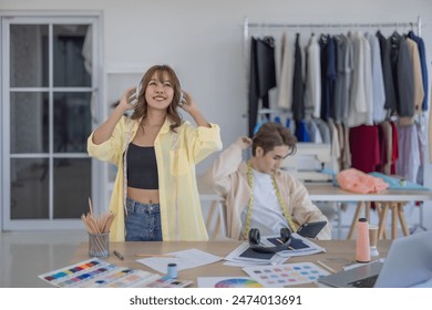 Two fashion designers enjoying creative workspace, surrounded by fabric samples, color palettes, clothing racks. One designer is listening to music with headphones while other is engrossed in tablet. - Powered by Shutterstock