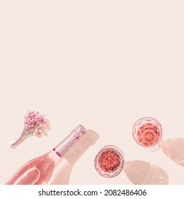 Two fashion glasses, bottle of rose wine and small bouquet of flowers on pastel pink background with copy space. Summer drink concept, dark shadows. 