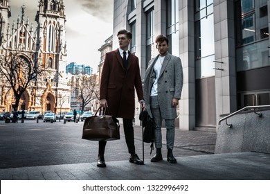 Two Fashion Men Models Posing On City Street. Wearing In Classic And Casual Closes. Suit, Coat, Shirt, Sweater, Boots And Leather Bag. Men Model Test Near Business Center