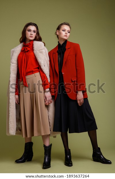 Two fashion models in black short \
pants, beige coat, long skirt,  bow blouse. Beautiful young women.\
Studio shot, Full length portrait. Green background.\
