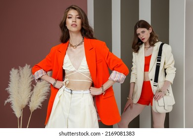 Two Fashion Models In Orange And Cream White Looks. Jacket, Blazer, Shorts, Top, Handbag. Beautiful Young Women. Rosy Brown And Striped Corrugated Wall, Dry Branches. Asian And European Girls