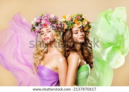 Two Fashion Women in Flower Wreath. Brunette and Blonde Beauty Models with Curly Hairstyle in Pink and Green Chiffon Dress with Flying Fabrics dreamy enjoying Spring Blossom over Yellow