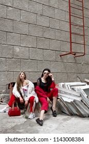 Two Fashion Young Girl Siting On The Backyard. Stylish Woman In A Red Suit And Sunglasses And In White Fur And Sport Red Pants .Many Old Demontaged Windows And Red Ladder.