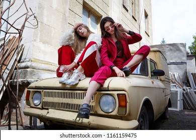 Two Fashion Young Girl Siting On The Backyard. Stylish Woman In A Red Suit And Sunglasses And In White Fur And Sport Red Pants .Emotioanl Shot.Motion.