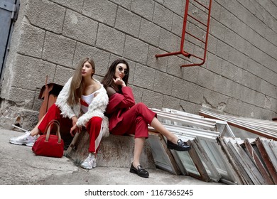 Two Fashion Young Girl Siting On The Backyard. Stylish Woman In A Red Suit And Sunglasses And In White Fur And Sport Red Pants .Many Old Demontaged Windows And Red Ladder.