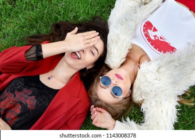 Two Fashion Young Happy Girl Laying On Green Grass .Laughing And Show Tongue The Other In Blue Sunglases.Stylish Woman In Red Jacket And Other In White Fur And T-shirt.Blue Sunglasses. .Fashion