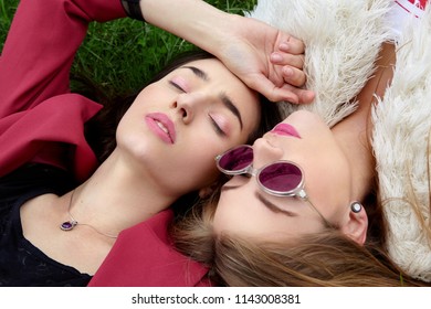 Two Fashion Young Happy Girl Laying On Green Grass .Close Eyes And Other In Blue Sunglases.Stylish Woman In Red Jacket And Other In White Fur And T-shirt.Pink Sunglasses.Fashion.World In Pink Glasses