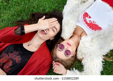 Two Fashion Young Happy Girl Laying On Green Grass .Laughing And Show Tongue The Other In Pink Sunglases.Stylish Woman In Red Jacket And Other In White Fur And T-shirt.Pink Sunglasses. 