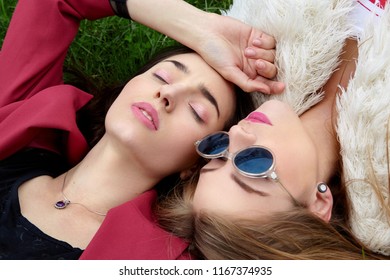 Two Fashion Young Happy Girl Laying On Green Grass .Close Eyes And Other In Blue Sunglases.Stylish Woman In Red Jacket And Other In White Fur And T-shirt.Blue Sunglasses. .Fashion