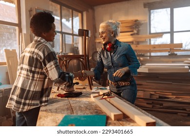 Two fashionable female carpenters are in their  workshop shaking hands and restoring furniture.