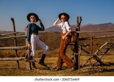 Two fashionable models wearing stylish autumn outfits with trendy hats, sunglasses, bags, cowboy boots, posing in mountains. Full length outdoor portrait. Copy, empty space for text