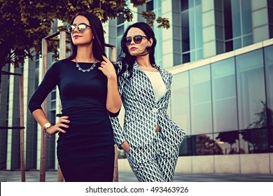 Two fashionable women in stylish clothes and sunglasses posing in a middle of business urban district.