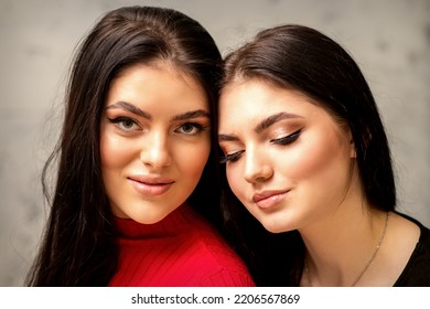 Two Fashionable Young Women. Portrait Of The Two Beautiful Female Models With Long Hair And Makeup. Two Beauty Young Caucasian Women With A Black Hairstyle On The Background Of A Gray Wall