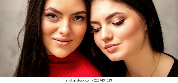 Two Fashionable Young Women. Portrait Of The Two Beautiful Female Models With Long Hair And Makeup. Two Beauty Young Caucasian Women With A Black Hairstyle On The Background Of A Gray Wall