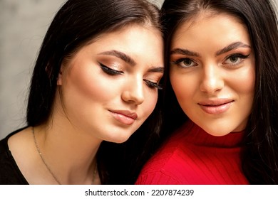 Two Fashionable Young Women. Portrait Of The Two Beautiful Female Models With Long Hair And Makeup. Two Beauty Young Caucasian Women With A Black Hairstyle On The Background Of A Gray Wall