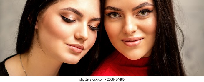 Two Fashionable Young Women. Portrait Of The Two Beautiful Female Models With Long Hair And Makeup. Two Beauty Young Caucasian Women With A Black Hairstyle On The Background Of A Gray Wall