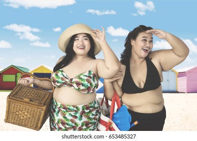 Two Fat Women Looking Something Near Cottage While Carrying Picnic Basket And Beach Items 