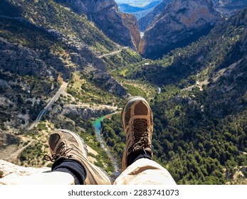 Two feet in hiking boots dangling in a ravine. In the background a valley with mountains and river.