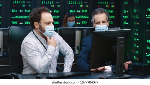 Two fellow programmers sit in office at desk in medical masks and discuss workflow looking at computer.