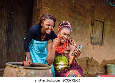 Two female Africans happily looking into the smart phone outside a village mud house that have a big water calabash