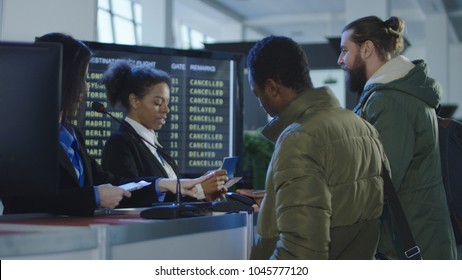 Two Female Airport Security Personnel Checking Identification At A Check-in Or Boarding Counter At The Departure Terminal Holding The Passports Of Two Male Passengers.