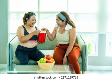 Two Female Asian Oversized Friends Wearing Spores Bras Waiting To Work Out Sit On The Couch Chatting To Eat Popcorn Junk Food In The Spores Club Living Room. Weight Loss Concept