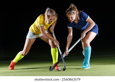 Two female athletes engage in a heated hockey competition under bright lights, showcasing their skills and determination on the field. The atmosphere is charged with competitive spirit. - Powered by Shutterstock