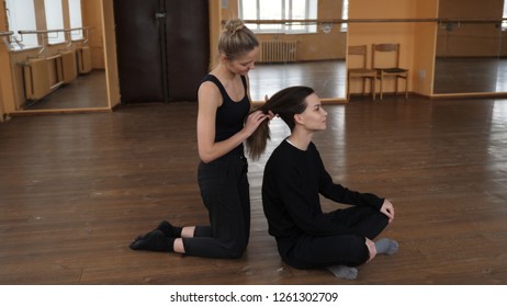 Two Female Blonde And Brunette Ballet Dancers Wearing Tights Stretching In A Studio And One Tying The Others Hair Into A Low Pony Tail
