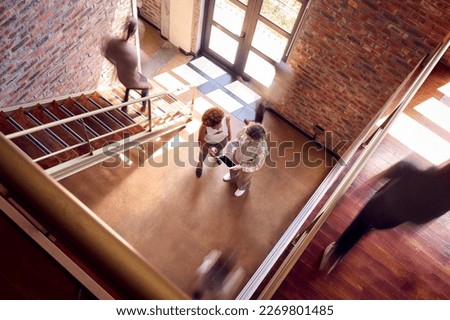 Two Female Business Colleagues With Digital Tablet Having Informal Meeting By Stairs In Busy Office