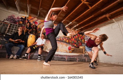 Two female capoeira performers sparring indoors while music plays