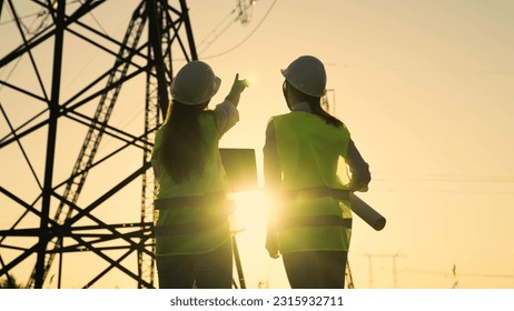 Two Female civil engineers work together using computer on power line. Joint work of power engineers with computer in protective helmets, maintenance of power lines in outdoors. Green energy.