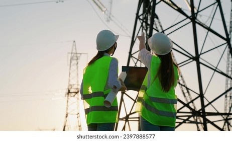 Two Female civil engineers work together using computer on power line. Joint work of power engineers with computer in protective helmets, maintenance of power lines in outdoors. Green energy. - Powered by Shutterstock