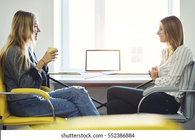 Two female colleagues discussing ideas of planning startup project of marketing company during friendly atmosphere with cup of coffee sitting near modern laptop computer with mock up screen in office