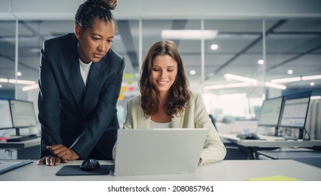 Two Female Colleagues Fondly Discussing Work, Smile while Showing Project on Laptop in Diverse Modern Business Office. Experienced Manager and Young Employee Discuss a Analytical Data.