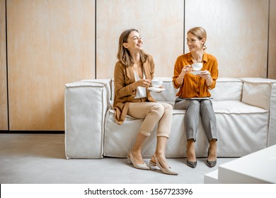 Two female colleagues having fun conversation, sitting together on the couch during a coffee break in the office