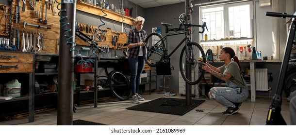 Two Female Cycling Workers Fixing Or Installing Wheels On Bicycle In Workshop. Young Multiethnic Women Colleaugues. Bike Service, Repair And Upgrade. Garage Interior With Tools And Equipment