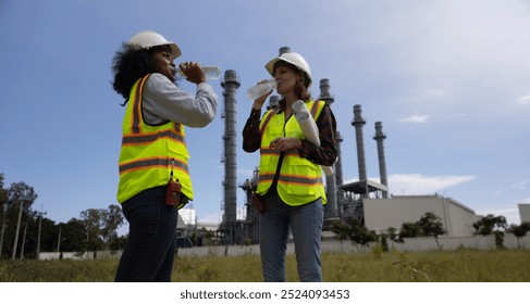 Two female engineers, from different backgrounds, take a break to drink water while working at a thermal power plant site.