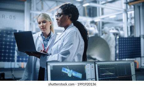 Two Female Engineers Talk, Use Computer Working At Aerospace Satellite Manufacturing Facility. Diverse Team Of Multi-Ethnic Scientists Doing Science Technology Research For Space Exploration Program