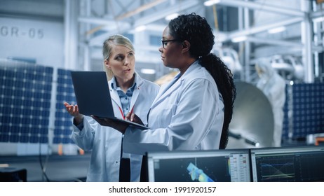 Two Female Engineers Talk, Use Computer Working At Aerospace Satellite Manufacturing Facility. Diverse Team Of Multi-Ethnic Scientists Doing Science Technology Research For Space Exploration Program