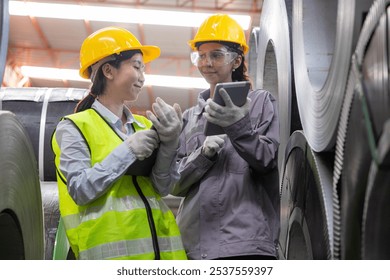 Two female engineers wearing safety gear inspect large metal coils in a manufacturing plant, discussing quality and production processes. - Powered by Shutterstock