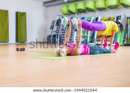 Two female fitness models doing yoga exercises, one lying on floor mat holding legs of another above her in a sports center