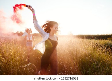 Two Female Friends Camping At Music Festival Running Through Field With Smoke Flare