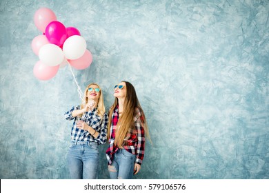 Two female friends in checkered shirts having fun with air balloons on the blue wall background