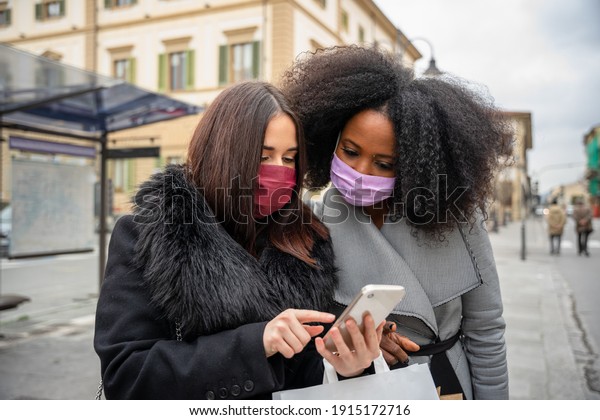 Two female friends in city at bus stop after\
shopping for sales check smartphone while looking for a taxi\
service or shared car transportation during Coronavirus Covid-19\
pandemic wearing face masks