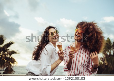 Two female friends enjoying ice cream together on a summer day. Cheerful young women eating icecream at seaside promenade.