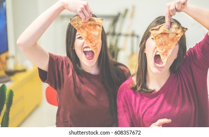 Two Female Friends Holding Pizza Slices In Front Of Their Faces - Young People Having Fun Eating Dinner - Rebel Concept - Focus On Left Slice - Warm Cinematic Filter