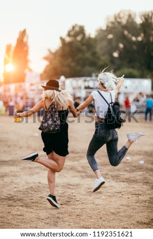 Two female friends jumping and having fun at music festival. Back view