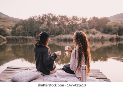 Two female friends in knitted warm sweaters having picnic near lake with autumn forest and lake on the background. Cozy fall atmosphere. - Powered by Shutterstock