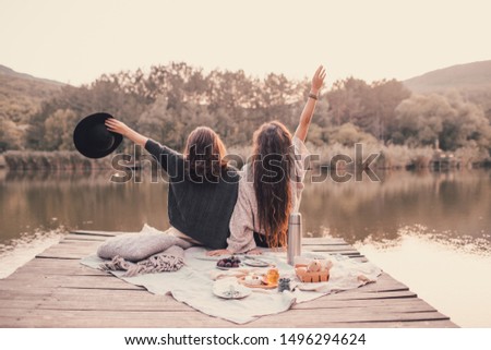 Two female friends in knitted warm sweaters having picnic near lake with autumn forest and lake on the background. Cozy fall atmosphere.